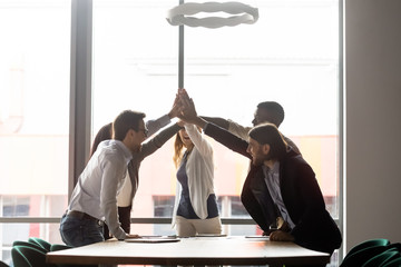 Excited diverse businesspeople give high five engaged in teambuilding activity at office briefing, overjoyed multiracial colleagues join hands show support and loyalty, motivated for shared goal