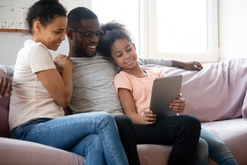 Wall Mural - Happy african american couple looking at tablet screen with daughter. Diverse smiling family of mother, father and girl sitting on couch using mobile device and gadget together.