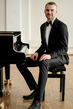 Portrait Of Young Pianist In Formal Elegant Suit With Bow Tie, Sitting Next To Classical Instrument Piano And Looking At Camera