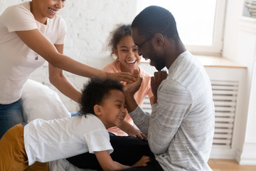 Wall Mural - Happy african american family tickle children and having good time on bed. Young diverse smiling husband and wife with cute daughter and son sitting in bedroom at home playing with kids.