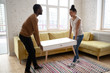 © fizkes - Young happy african american couple moving wooden table. Attractive family of diverse woman and man relocation furniture in new flat. Husband and wife perform permutation. First time buyers
