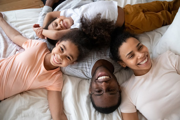 Wall Mural - Happy african american parents with little children lying on bed top view, looking at camera, smiling beautiful mother and father enjoying free time with cute adorable daughter and son.