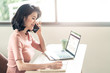© Kawee - Asian young business woman sit and write note in a notebook while talk on phone and open files in laptop. During Coronavirus crisis Covid19 outbreak girl work from home for prevent contract a disease.