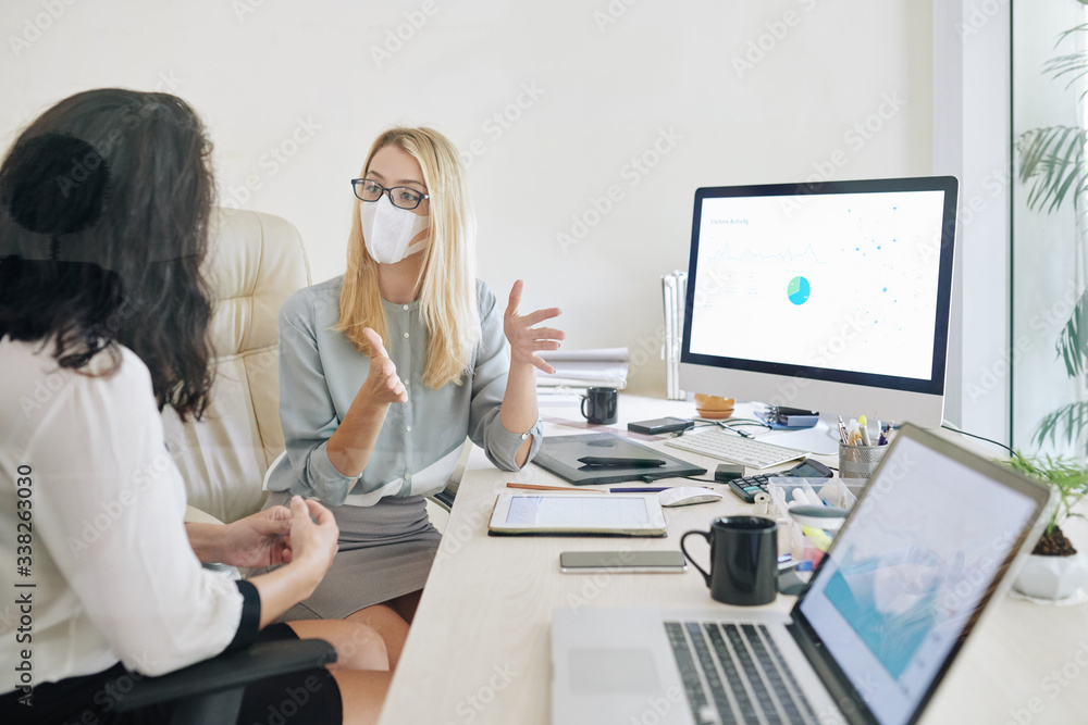 Young businesswoman in glasses and medical mask actively gesturing when explaining idea to collesgue - obrazy, fototapety, plakaty 