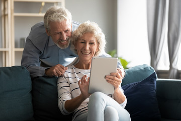 Wall Mural - Happy older couple using computer tablet together at home, excited mature man and woman looking at mobile device screen, shopping or chatting online, sitting on cozy couch in living room