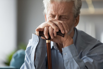 Close up sad tired older man holding hands on walking stick, problem with health, upset unhappy mature senior male using wooden cane during rehabilitation, elderly people healthcare