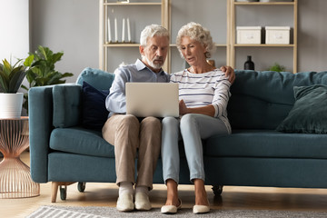 Wall Mural - Mature man and woman using laptop together, looking at screen, older spouses browsing apps, shopping or chatting online, making video call, using bank service, sitting on cozy sofa in living room