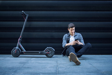 Shot of young caucasian hipster using smart phone while siting on the ground next to his electric scooter against black wall background of modern business building.