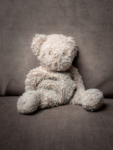A Tired, Old Teddy Bear Sitting, Collapsed On A Grey Cushion