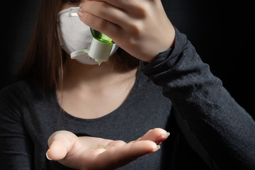 Wall Mural - Coronavirus protection. Young woman in a white antibacterial medical mask smears her hands with antiseptic gel on a black background