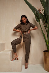 Wall Mural - Beautiful woman fashion model brunette hair tanned skin wear green overalls button suit sandals high heels accessory clothes style journey safari summer collection plant flowerpot wall stairs.