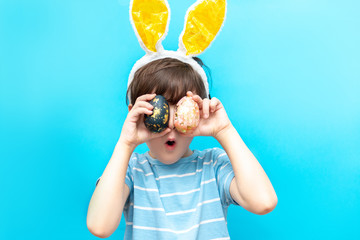 little boy with hare ears holds two painted eggs on a blue background