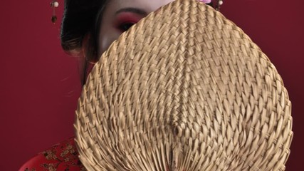 Poster - A close-up view of a beautiful young geisha woman in traditional japanese kimono is posing with wooden hand fan while moving it from one side to another isolated over red background