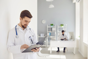 Wall Mural - A male doctor with a stethoscope reads a clipboard while standing in a clinic.