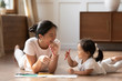 © fizkes - Educational pastime develop creativity skill in kid concept. Asian mother her small daughter lying on warm wooden floor in sunny cozy living room, mom teach girl paint use album and colourful pencils