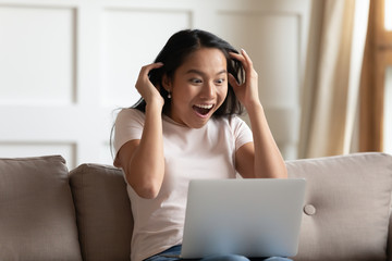 Crazy excited asian woman sit on couch read news online on laptop feels shocked amazed and surprised. Unbelievable sell-out web-store discounts, on-line lottery win, new free cool application concept