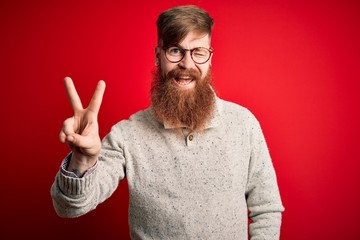 Handsome Irish redhead man with beard wearing casual sweater and glasses over red background smiling with happy face winking at the camera doing victory sign with fingers. Number two.