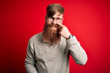 Canvas Print - Handsome Irish redhead man with beard wearing casual sweater and glasses over red background Pointing with hand finger to face and nose, smiling cheerful. Beauty concept