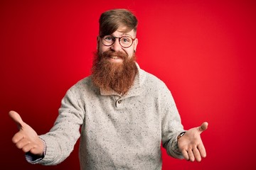 Handsome Irish redhead man with beard wearing casual sweater and glasses over red background smiling cheerful with open arms as friendly welcome, positive and confident greetings