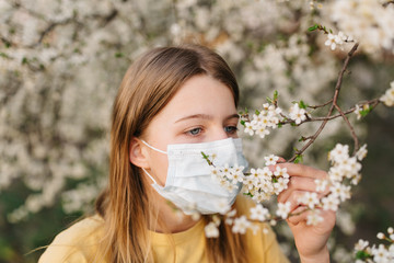 portrait of sad young woman in protective medical face mask with flowers near blooming tree in spring time. Coronavirus protection. epidemic of coronavirus. the aroma a tree in the garden on a spring