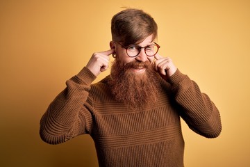 Canvas Print - Handsome Irish redhead man with beard wearing glasses and winter sweater over yellow background covering ears with fingers with annoyed expression for the noise of loud music. Deaf concept.