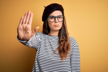 Wall Mural - Young beautiful brunette woman wearing french beret and glasses over yellow background doing stop sing with palm of the hand. Warning expression with negative and serious gesture on the face.