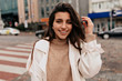 © PhotoBook - Close up outside portrait of smiling pretty woman with dark hair wearing beige sweater and white coat walking on the street with happy emotions on background of old buildings