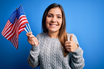 Young patriotic woman holding usa flag on independence day 4th of july over blue background happy with big smile doing ok sign, thumb up with fingers, excellent sign