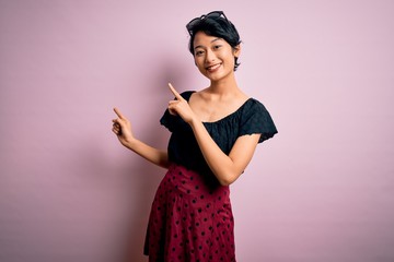 Young beautiful asian girl wearing casual dress standing over isolated pink background smiling and looking at the camera pointing with two hands and fingers to the side.