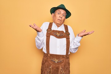 Sticker - Senior grey-haired man wearing german traditional octoberfest suit over yellow background clueless and confused expression with arms and hands raised. Doubt concept.