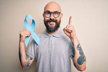 Poster - Handsome bald man with beard and tattoos holding blue cancer ribbon over isolated background surprised with an idea or question pointing finger with happy face, number one