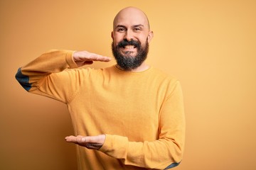 Poster - Handsome bald man with beard wearing casual sweater standing over yellow background gesturing with hands showing big and large size sign, measure symbol. Smiling looking at the camera. Measuring