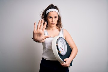 Poster - Young beautiful woman with curly hair holding weighing machine over white background doing stop sing with palm of the hand. Warning expression with negative and serious gesture on the face.