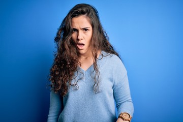 Young beautiful woman with curly hair wearing blue casual sweater over isolated background In shock face, looking skeptical and sarcastic, surprised with open mouth