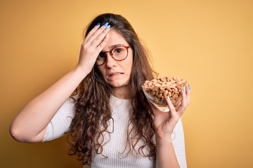 Sticker - Beautiful woman with curly hair holding bowl with healthy peanuts over yellow background stressed with hand on head, shocked with shame and surprise face, angry and frustrated. Fear and upset