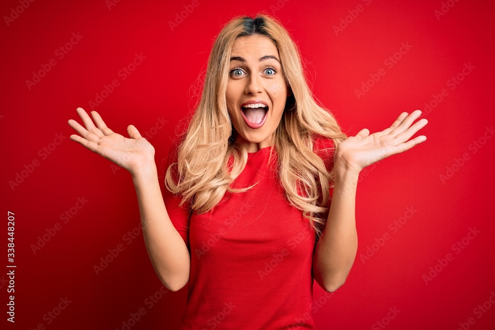 Young beautiful blonde woman wearing casual t-shirt standing over isolated red background celebrating crazy and amazed for success with arms raised and open eyes screaming excited. Winner concept - obrazy, fototapety, plakaty 