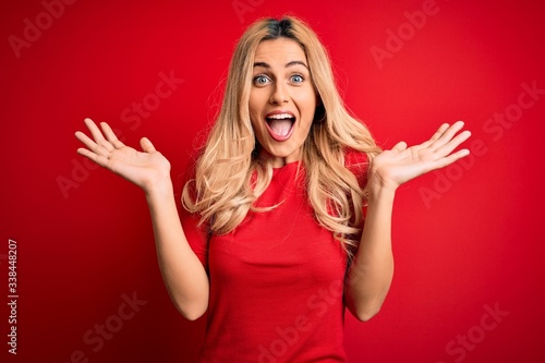 Fotografía Young beautiful blonde woman wearing casual t-shirt standing over isolated red background celebrating crazy and amazed for success with arms raised and open eyes screaming excited