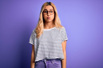 Poster - Young beautiful blonde woman wearing striped t-shirt and glasses over purple background puffing cheeks with funny face. Mouth inflated with air, crazy expression.