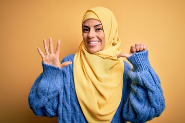 Wall Mural - Young beautiful brunette muslim woman wearing arab hijab over isolated yellow background showing and pointing up with fingers number six while smiling confident and happy.