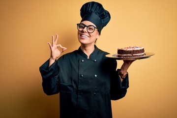 Poster - Young beautiful brunette baker woman wearing cooker uniform and hat holding cake doing ok sign with fingers, excellent symbol