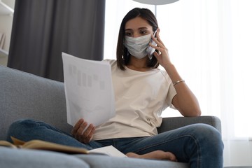 Canvas Print - Young woman wearing protective mask in quarantine and using smartphone while sitting on comfortable sofa