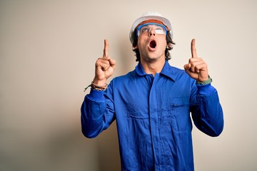 Young constructor man wearing uniform and security helmet over isolated white background amazed and surprised looking up and pointing with fingers and raised arms.