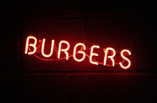 A Vintage Retro American Style Neon Sign Advertising Burgers. A Dark Setting With Neon Tube Lighting Up The Dark In Red. Food Advertising And Signage In The Food Industry