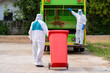 © visoot - Two garbage men in hazmat PPE protective clothing wear medical rubber gloves working together on emptying dustbins for trash removal with truck loading waste and trash bin,Coronavirus Disease 2019.