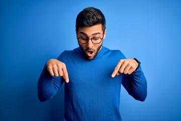young handsome man with beard wearing casual sweater and glasses over blue background pointing down 