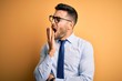 © Krakenimages.com - Young handsome businessman wearing tie and glasses standing over yellow background bored yawning tired covering mouth with hand. Restless and sleepiness.