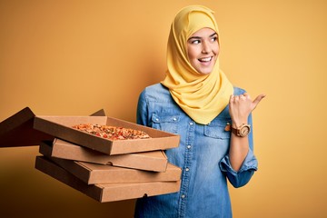 Sticker - Young girl wearing muslim hijab holding boxes with Italian pizza over yellow background pointing and showing with thumb up to the side with happy face smiling