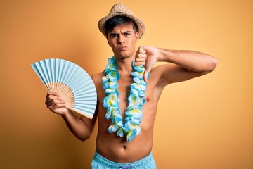 Poster - Young tourist man on vacation wearing swimwear and hawaiian lei flowers holding hand fan with angry face, negative sign showing dislike with thumbs down, rejection concept