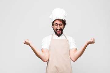 young crazy chef looking extremely happy and surprised, celebrating success, shouting and jumping against white wall