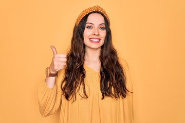 Wall Mural - Young beautiful woman wearing casual t-shirt and diadem standing over yellow background doing happy thumbs up gesture with hand. Approving expression looking at the camera showing success.